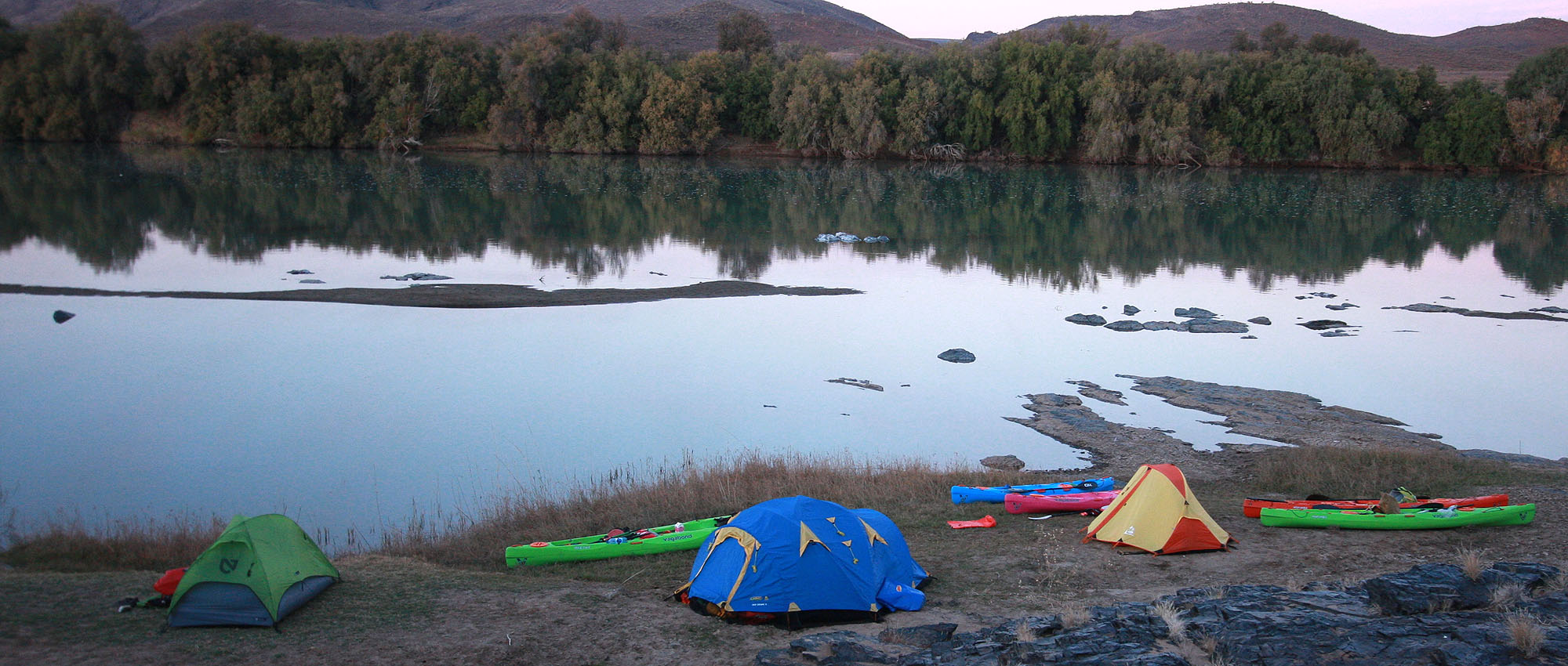 Tents and kayaks on a rocky riverbank at dusk.